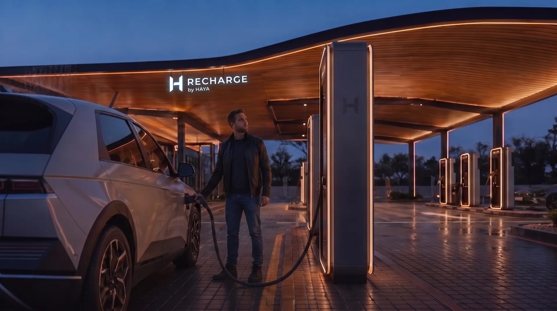 A driver charging his SUV under a warm wooden canopy with the RECHARGE by HAYA logo illuminated at dusk